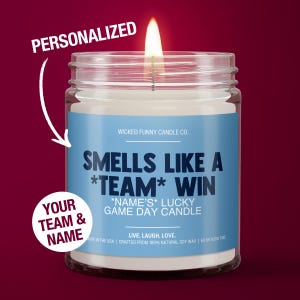 May include: A personalized candle with a blue label that says "Smells Like A *TEAM* WIN *NAME'S* Lucky Game Day Candle". The candle is in a clear glass jar with a white lid. The label also says "Wicked Funny Candle Co.", "Live, Laugh, Love.", "Made in the USA", "Crafted from 100% Natural Soy Wax", and "60 hr Burn Time".