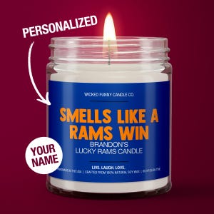 May include: A personalized candle with a blue and orange label that reads "Smells Like A Rams Win" and "Brandon's Lucky Rams Candle". The candle is in a clear glass jar with a white wax interior and a lit wick.