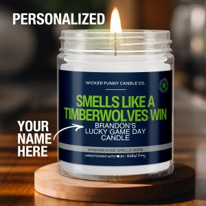 May include: A personalized candle with a blue and green label that reads "Smells Like A Timberwolves Win" and "Brandon's Lucky Game Day Candle". The candle is in a clear glass jar with a white wax interior and a lit wick.
