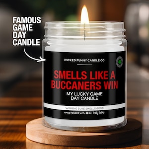 May include: A lit glass jar candle with a black label that reads "Smells Like a Buccaneers Win." The candle is on a wooden coaster. Text on the label includes "My Lucky Game Day Candle" and "Winning Sure Smells Good."