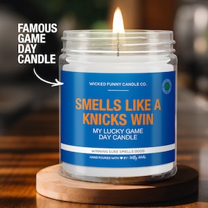 May include: A clear glass candle jar with a blue and orange label that reads "Smells Like A Knicks Win" and "My Lucky Game Day Candle". The candle is lit and the wick is visible.
