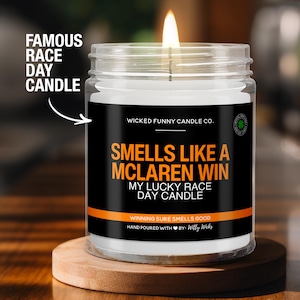 May include: A black and orange candle with the text "Smells Like A McLaren Win" and "My Lucky Race Day Candle". The candle is in a clear glass jar with a black label and a green shamrock logo.
