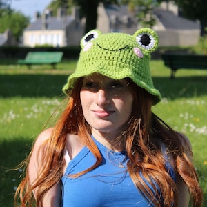 Puede incluir: Una mujer con el pelo largo y rojo lleva un sombrero de rana de ganchillo verde con ojos negros y mejillas rosas. El sombrero tiene un ala ancha y está puesto en su cabeza. Lleva una camisa azul y está de pie en un campo de hierba.