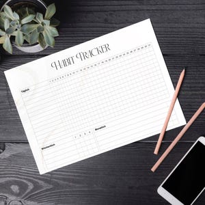 Puede incluir: Un rastreador de hábitos blanco con el título "Habit Tracker" en negro sobre una superficie de madera negra. El rastreador tiene una cuadrícula para 31 días y 4 semanas. Hay dos lápices y un teléfono inteligente en la superficie.