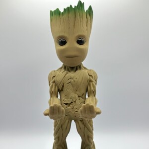 May include: A brown and green figurine of a character from the Guardians of the Galaxy movie franchise. The character is standing with its arms outstretched and has a serious expression on its face. The figurine is on a black base.