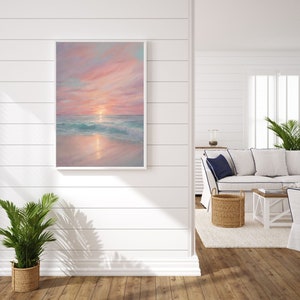 Puede incluir: Una pintura enmarcada de una puesta de sol rosa y naranja sobre una playa con agua turquesa. La pintura est&aacute; colgada en una pared blanca en una habitaci&oacute;n con un sof&aacute; blanco y una cesta de mimbre.