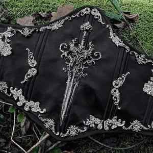 May include: Black corset with ornate silver embellishments and a central sword design. The corset features detailed scrollwork and decorative accents along the edges, creating a gothic or fantasy aesthetic.