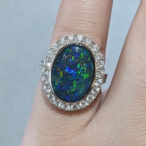 May include: Oval-shaped ring featuring a large, iridescent black opal stone surrounded by a halo of small, clear gemstones. The opal displays vibrant flashes of blue, green, and gold. The ring is set in a silver-colored metal.