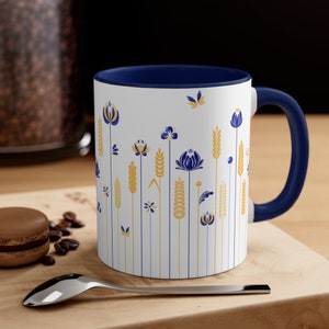 May include: A white ceramic mug with a blue rim and handle. The mug features a repeating pattern of stylized flowers and wheat stalks in shades of blue and gold.