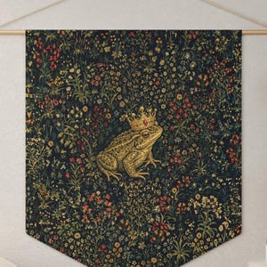 May include: A wall hanging banner featuring a dark navy blue background with a dense, colorful floral pattern and a central illustration of a golden toad wearing a crown. The banner is suspended from a wooden rod with a thin string for display.