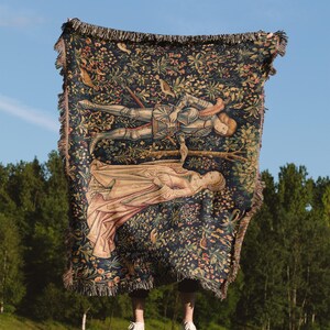 May include: A woven throw blanket featuring a dark blue background with a dense floral pattern and a central illustration of a knight in armor holding hands with a figure in a long gown. The blanket has a fringed edge on all four sides.