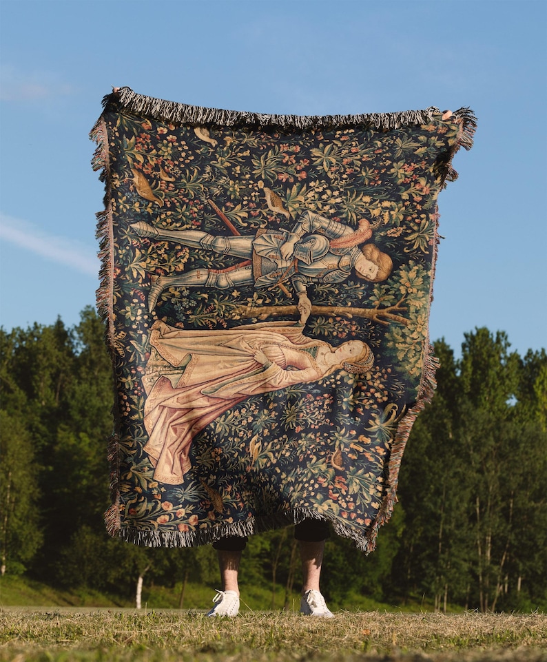 May include: A woven throw blanket featuring a dark blue background with a dense floral pattern and a central illustration of a knight in armor holding hands with a figure in a long gown. The blanket has a fringed edge on all four sides.