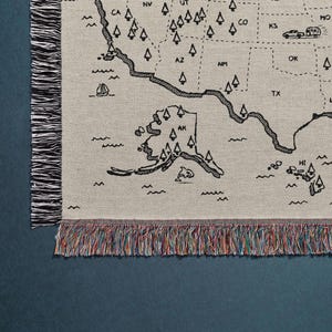 May include: A woven throw blanket featuring a black line-art map of the United States on a light beige background. The design includes state abbreviations, small tree icons, and travel illustrations like a camper and boat, finished with multicolored fringe.