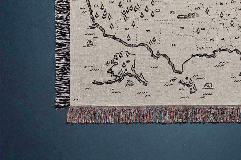 May include: A woven throw blanket featuring a black line-art map of the United States on a light beige background. The design includes state abbreviations, small tree icons, and travel illustrations like a camper and boat, finished with multicolored fringe.
