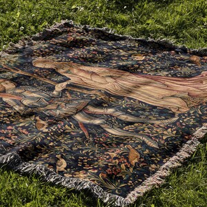 May include: A rectangular woven throw blanket featuring a dark blue background with a detailed scene of a figure in armor and a figure in flowing robes surrounded by colorful floral patterns and small birds, finished with a fringed edge.