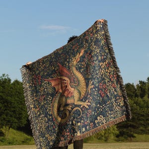 May include: A rectangular woven throw blanket featuring a dark blue background with a detailed illustration of a green dragon breathing fire, surrounded by colorful floral and botanical patterns, finished with a fringed edge on all four sides.