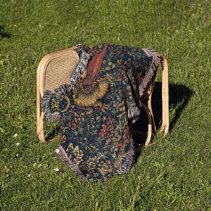 May include: A woven throw blanket featuring a dark blue background with a detailed pattern of a golden dragon, red flowers, and green foliage. The blanket has a fringed edge and is draped over a light-colored wooden chair with a cane back.