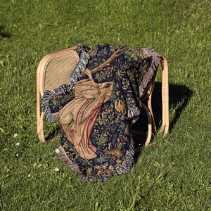 May include: A woven throw blanket featuring a dark blue background with a detailed botanical pattern of flowers, leaves, and a central figure in tan and brown tones. The rectangular blanket has a fringed edge and is draped over a light wood chair.