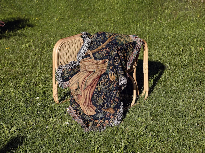 May include: A woven throw blanket featuring a dark blue background with a detailed botanical pattern of flowers, leaves, and a central figure in tan and brown tones. The rectangular blanket has a fringed edge and is draped over a light wood chair.