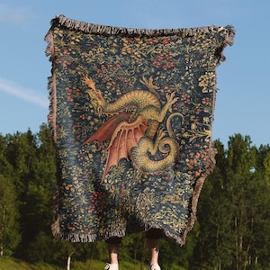 May include: A rectangular woven throw blanket featuring a dark blue background with a detailed pattern of colorful wildflowers and a large, central green dragon with red wings. The blanket has a fringed edge along all four sides of the fabric.