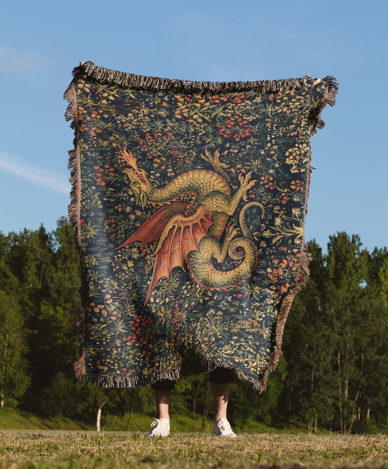 May include: A rectangular woven throw blanket featuring a dark blue background with a detailed pattern of colorful wildflowers and a large, central green dragon with red wings. The blanket has a fringed edge along all four sides of the fabric.