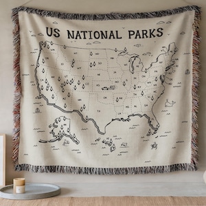May include: A cream-colored woven throw blanket featuring a black line-art map of the United States with the text US NATIONAL PARKS. The map includes state abbreviations, small tree icons, and a camper illustration, finished with multicolored fringe.