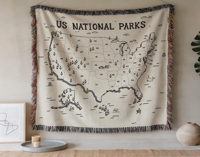 May include: A cream-colored woven throw blanket featuring a black line-art map of the United States with the text US NATIONAL PARKS. The map includes state abbreviations, small tree icons, and a camper illustration, finished with multicolored fringe.