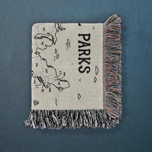 May include: A woven throw blanket in a light beige color featuring a black line-art map of the northeastern United States with the text PARKS printed vertically. The blanket has decorative multi-colored fringe along the right and bottom edges.