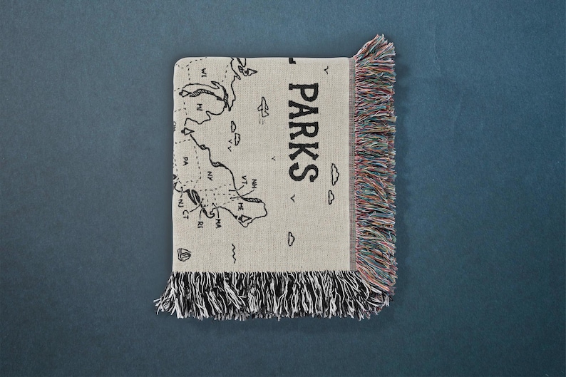 May include: A woven throw blanket in a light beige color featuring a black line-art map of the northeastern United States with the text PARKS printed vertically. The blanket has decorative multi-colored fringe along the right and bottom edges.