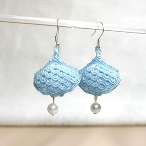 Peut inclure: Une paire de boucles d'oreilles en macramé bleu clair en forme de cloche. Chaque boucle d'oreille est ornée d'une petite perle blanche ronde suspendue en bas. Les boucles sont suspendues à des crochets d'oreilles argentés.