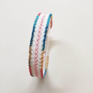 Può includere: Un braccialetto intrecciato colorato con un motivo a spina di pesce. Il braccialetto presenta una base bianca con accenti rosa, blu, rossi e dorati. Il braccialetto è realizzato in materiale intrecciato ed è progettato per essere indossato al polso.