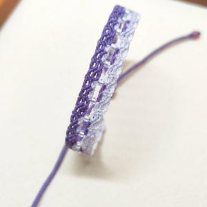 Peut inclure: Un bracelet en macramé aux tons violets et blancs. Le bracelet présente un motif tissé avec une fermeture coulissante. Le bracelet est fait d'un matériau souple et flexible et convient à un usage quotidien.