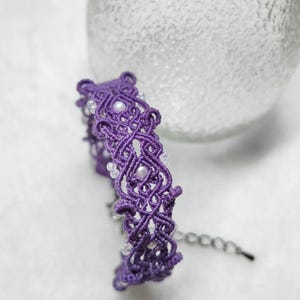 Op de afbeelding: Een paarse macramé armband met witte kralen en een zilveren kettingsluiting.