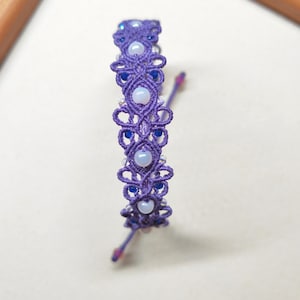Peut inclure: Un bracelet macramé artisanal aux tons violets, avec un travail de nœuds complexe et orné de petites perles rondes blanches et bleues. Le bracelet est conçu avec un motif délicat et une fermeture coulissante.