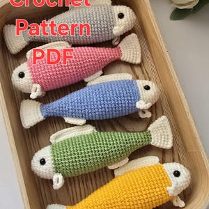 May include: A wooden tray holds five crocheted fish in various colors: gray, pink, blue, green, and yellow. Each fish has a white belly and tail fin, with black bead eyes. The text "Fish Crochet Pattern PDF" is displayed at the top.