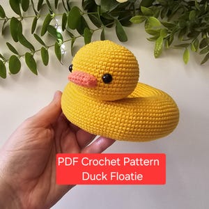 May include: A handmade yellow crochet duck floatie with a round head, a pink beak, and black eyes. The duck floatie is designed with a circular body. Text on a red rectangle reads "PDF Crochet Pattern Duck Floatle".