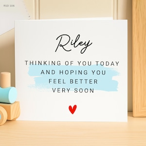 May include: A white card with the name "Riley" in cursive script. The card includes the message "Thinking of you today and hoping you feel better very soon" with a light blue brushstroke and a red heart.