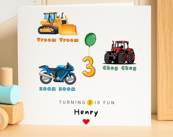 May include: White birthday card with illustrations of a yellow bulldozer, blue motorcycle, and red tractor. A green balloon is attached to a number 3. The text reads "Turning 3 is fun" and "Henry" with a red heart.
