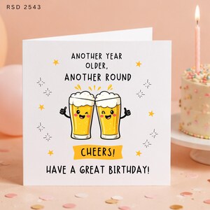 May include: A white square birthday greeting card featuring a cartoon illustration of two clinking beer glasses with faces. The text reads: ANOTHER YEAR OLDER, ANOTHER ROUND, CHEERS! HAVE A GREAT BIRTHDAY! The design includes yellow stars and sparkles.