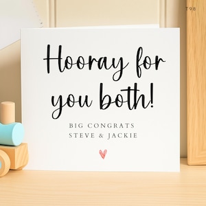 May include: A white greeting card with the words "Hooray for you both!" in black script. Below, it reads "BIG CONGRATS STEVE & JACKIE" with a small red heart. The card is on a wooden surface.