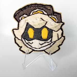 May include: Embroidered patch of a character with a black and yellow face, a white mask, and a brown hat. The patch is displayed on a clear acrylic stand against a white background. The patch has a detailed, textured appearance.