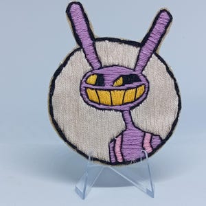May include: Embroidered patch featuring a purple cartoon character with a wide yellow grin and large ears. The character is set against a beige circular background with a black border. The patch is displayed on a clear stand.