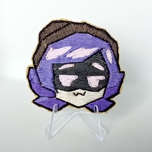 May include: Embroidered patch depicting a cartoon character with purple hair, a black mask, and a brown hat. The patch has a gold border and is displayed on a clear stand. The design is a stylized illustration.