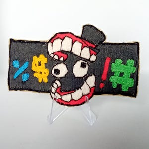 May include: Embroidered patch with a cartoon face, a wide mouth with red lining and white teeth. The face is black with white eyes and a small black hat. Symbols including a percent sign, a pound sign, an exclamation mark, and a hash are embroidered on a black background.