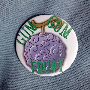 May include: A round white button with a purple fruit design and green text that reads "GUM GUM FRUIT". The fruit has a swirling pattern and a brown stem. The button is on a blue fabric background.