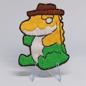 May include: Embroidered patch featuring a cartoon dinosaur wearing a brown cowboy hat. The dinosaur has a yellow body, white belly, and green legs. The patch is outlined in black.