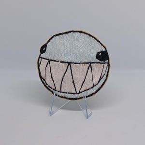 May include: A circular embroidered patch featuring a light blue upper section and a wide, pale pink mouth filled with sharp, black-outlined triangular teeth. The design includes two small black circular eyes and a thick black border outline.