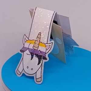 May include: A white unicorn bookmark with a glittery finish and a non-binary flag design. The unicorn has a gold horn. The bookmark is on a blue surface.