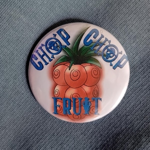 May include: A round white button with the words "CHOP CHOP" in blue, surrounding a cartoon image of a reddish-orange fruit with green leaves. The word "FRUIT" is written in blue at the bottom.