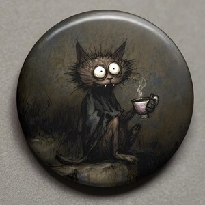 Puede incluir: Un botón redondo con una ilustración caprichosa de un gato con ojos grandes, sosteniendo una taza de té con vapor. El gato está representado en tonos oscuros, vistiendo una túnica oscura y sentado sobre una piedra. El fondo es una escena sombreada y apagada.